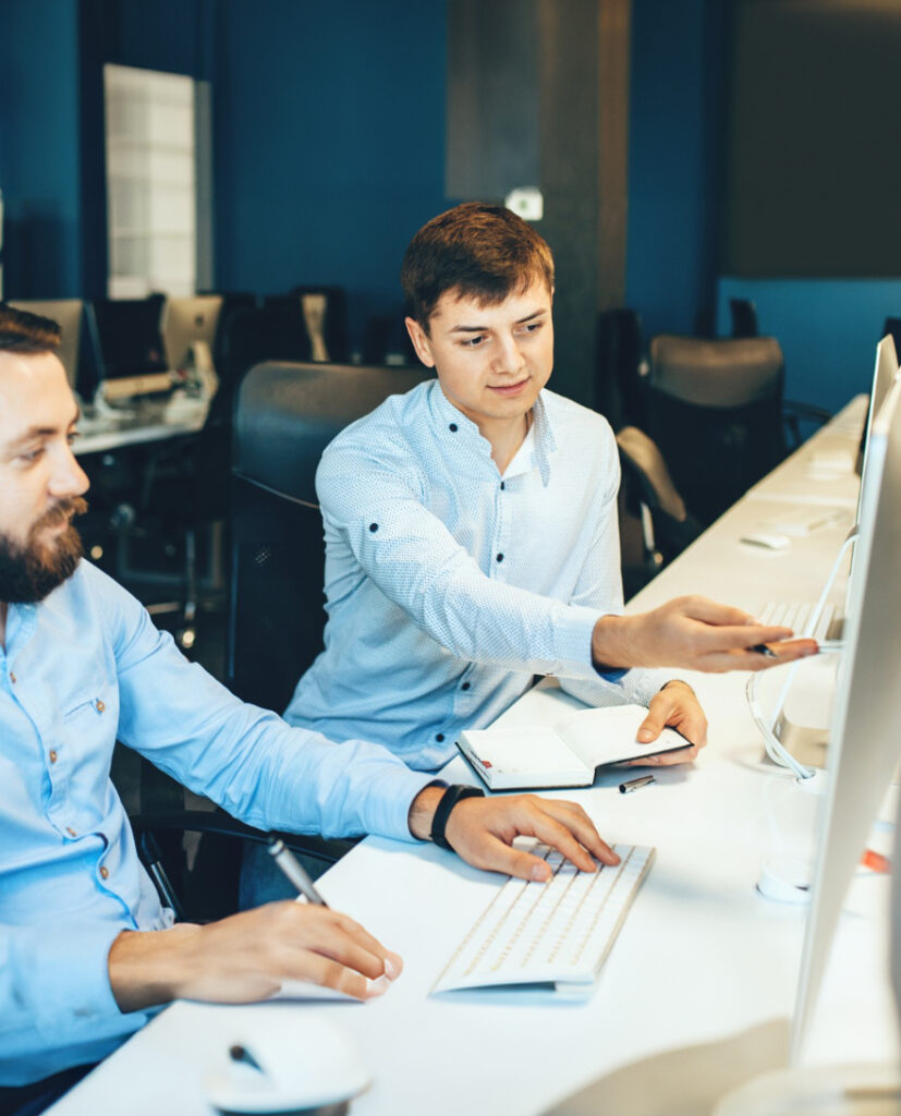 Two men in a modern office setting, one pointing at a computer screen while the other listens attentively, collaborating on a project.